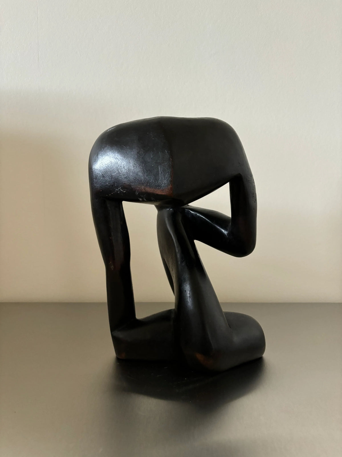 A Black Wooden Object - “The Thinker” by CU-RA-TED resembles a human figure bending forward with one knee on the floor, head resting on their thigh, and one arm bent, placing a hand on the ground. The piece showcases exquisite craftsmanship and minimalist design against a plain background.