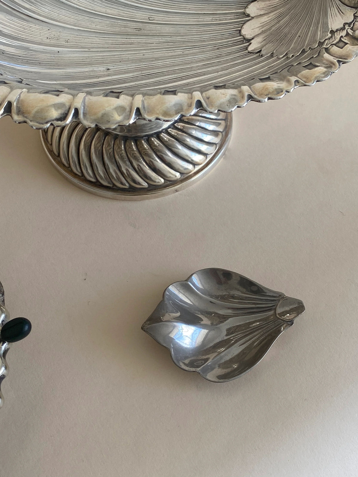 A close-up image of a decorative silver footed dish with a ribbed design and scalloped edges. Beside it is one of the vintage chrome leaf trays from the "Set 5 Vintage Chrome Leaf Trays" by Aesthete Label. Both items rest on a light-colored surface.