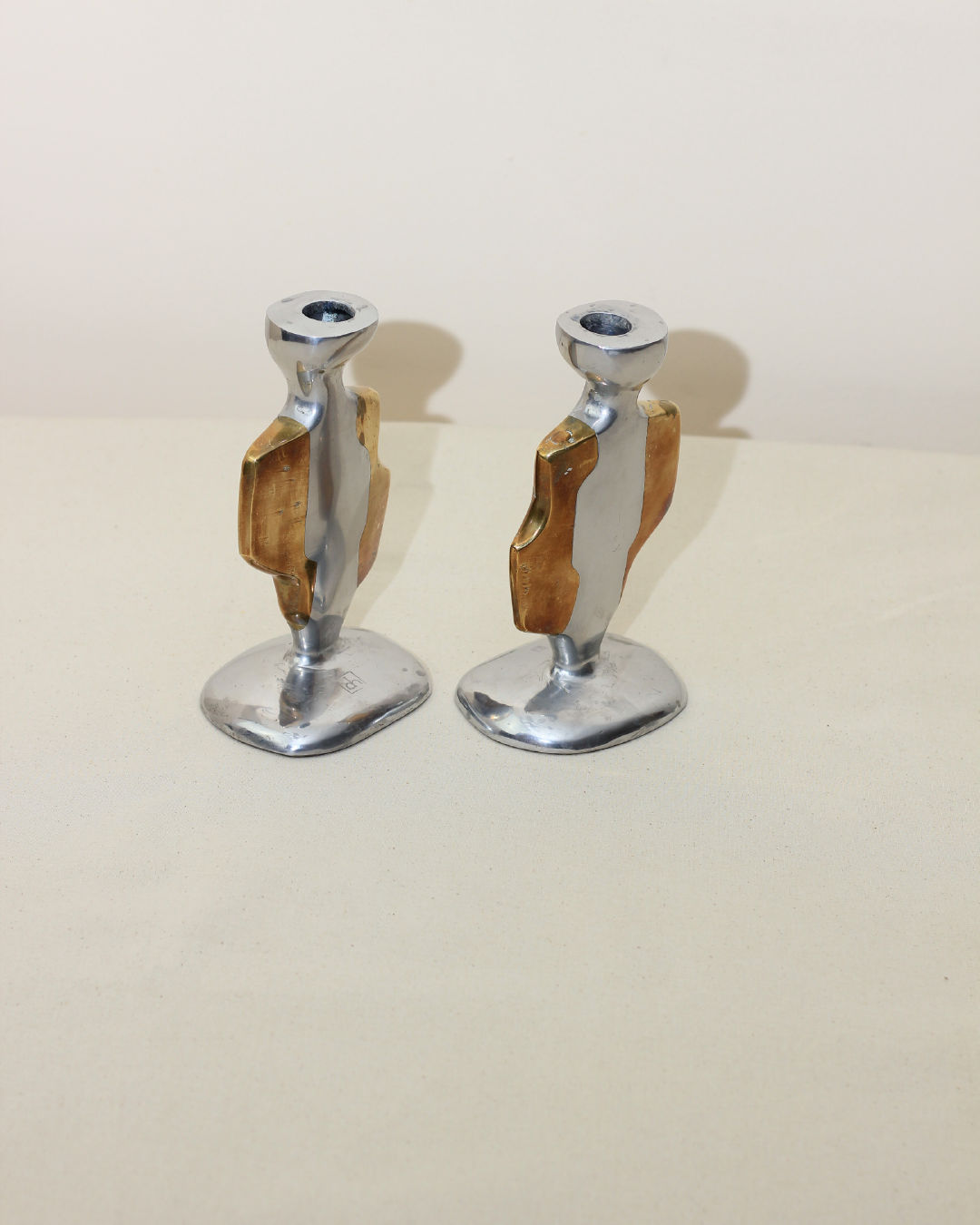 The Pair of Brutalist Candleholders by Boga Avante Shop features two metallic holders with abstract shapes in shiny silver and gold aluminum and brass, inspired by vintage David Marshall designs, displayed on a light surface with a cream background.
