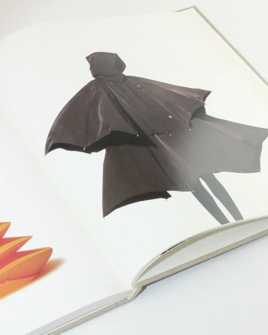 In the book "Irving Penn regards the work of Issey Miyake" from Boga Avante Shop, an open spread showcases a photograph of a person adorned in a dark, voluminous cloak-like garment reminiscent of Issey Miyake's designs. The minimalist image emphasizes the silhouette of the outfit against a light background, with an orange abstract shape partially visible on the left page.