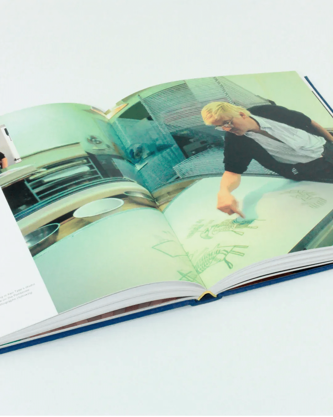 An open book displaying a photo of a chef in a kitchen, gesturing toward drawings on a whiteboard or table. The pages surrounding the photo reveal a kitchen setup with cooking utensils and equipment in the background, reminiscent of contemporary art that might be found in "That's the way I see it: David Hockney" by Boga Avante Shop.
