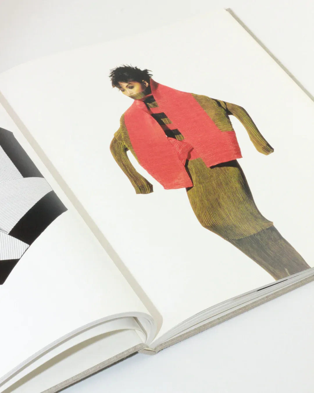 A photo of an open book showing an illustration of a person wearing a red scarf, depicted in a stylized manner with textured patterns reminiscent of Issey Miyake designs. The background is white, with part of another illustration visible on the left side, capturing the essence of fashion photography akin to Irving Penn's work. This scene perfectly encapsulates the spirit of "Irving Penn regards the work of Issey Miyake," available from Boga Avante Shop.