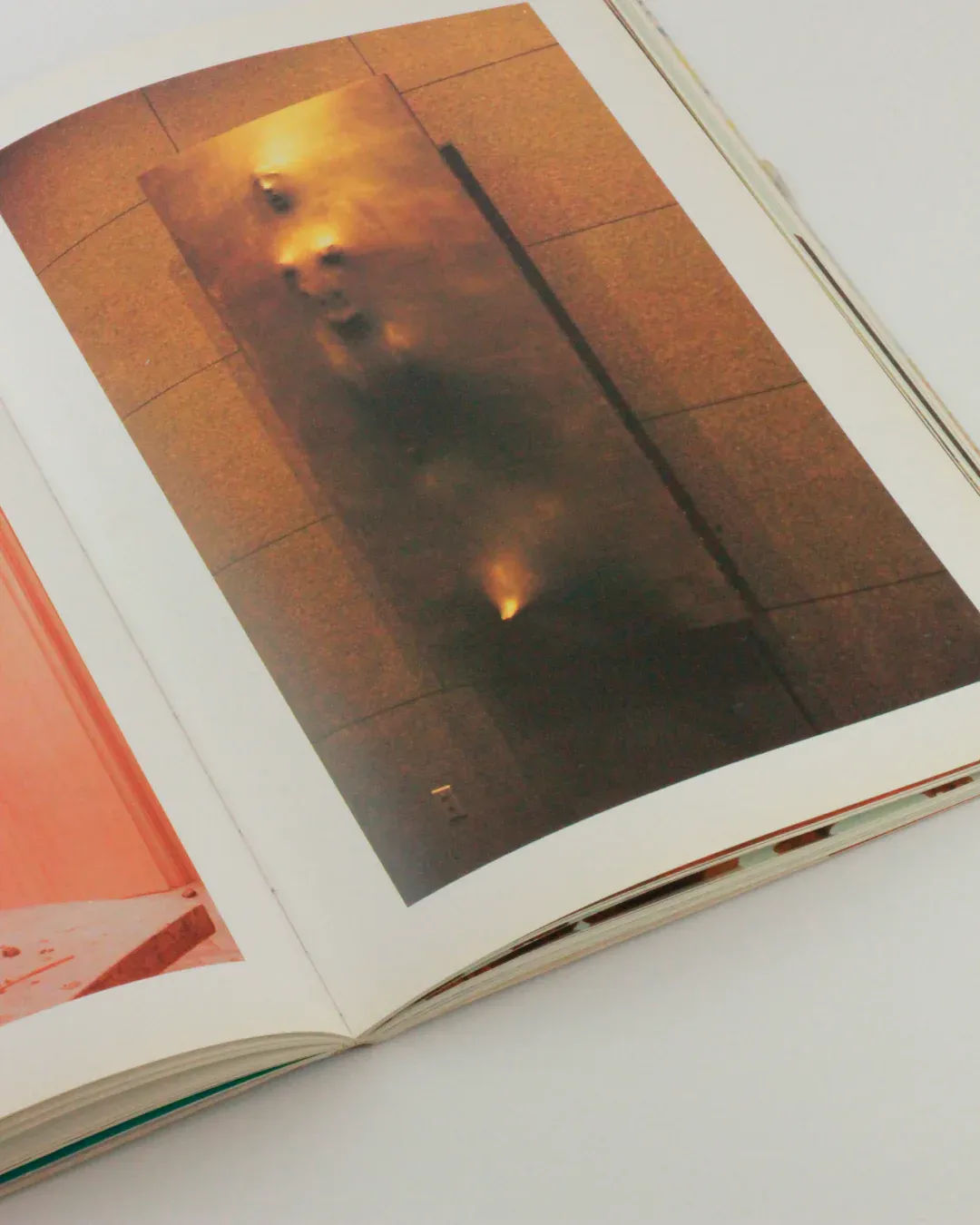 An Issey Miyake Vintage Book from Boga Avante Shop lies open, with the right page displaying a photograph of a wall adorned by an abstract, textured, dark and smoky area, likely part of an art installation. Evoking Issey Miyake's signature blend of modern technology and Japanese tradition, the left page reveals a glimpse of a pinkish hue in its small visible corner. The book is set against a plain backdrop.