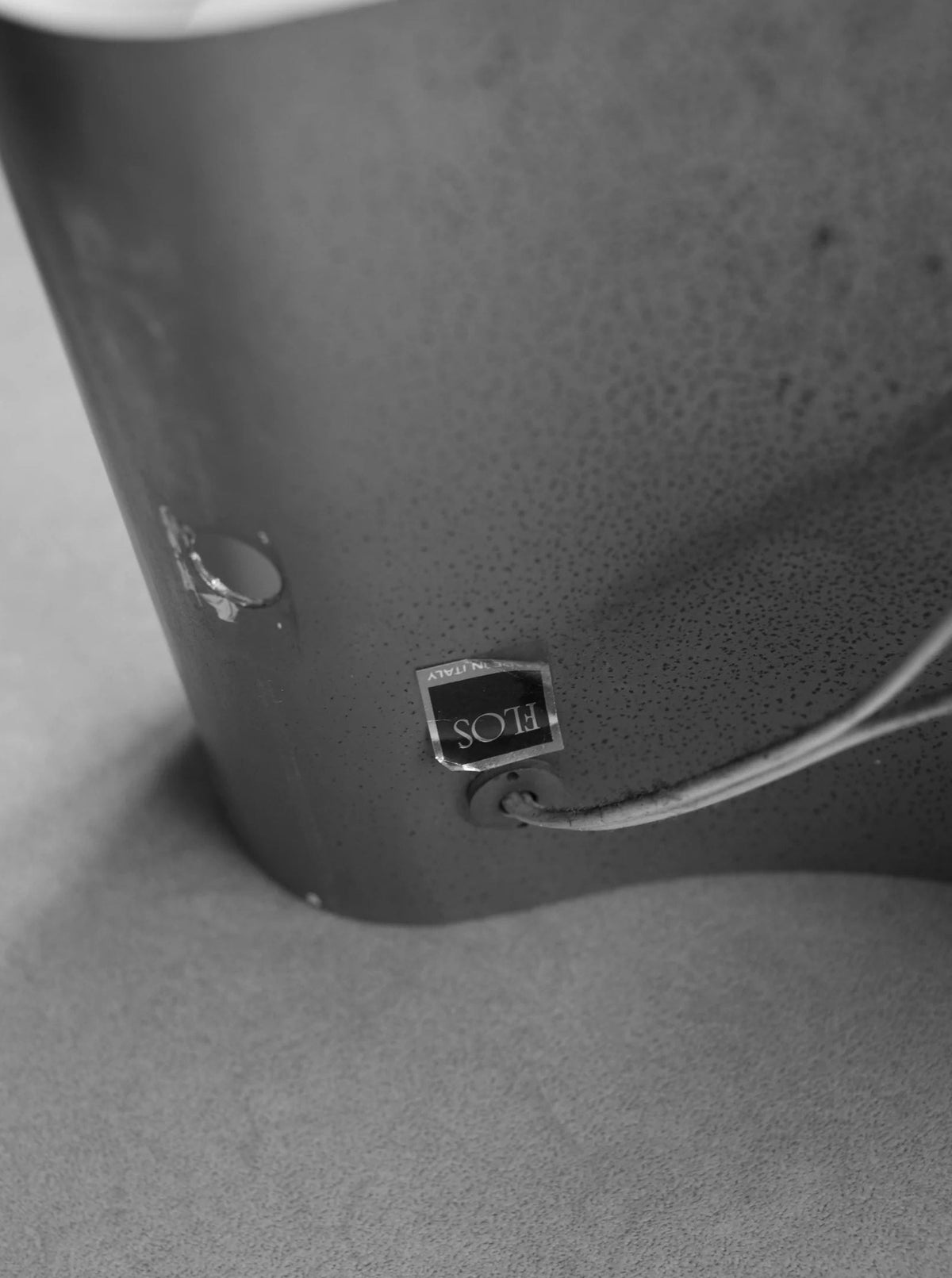Black and white image of the back of an object with a tag labeled "CU-RA-TED". The object, resembling a Chrome Wall Lamp by Tobia Scarpa 80's, appears to have a rough texture, and a cable is attached to it. The background is blurry, drawing focus to the object and the tag.