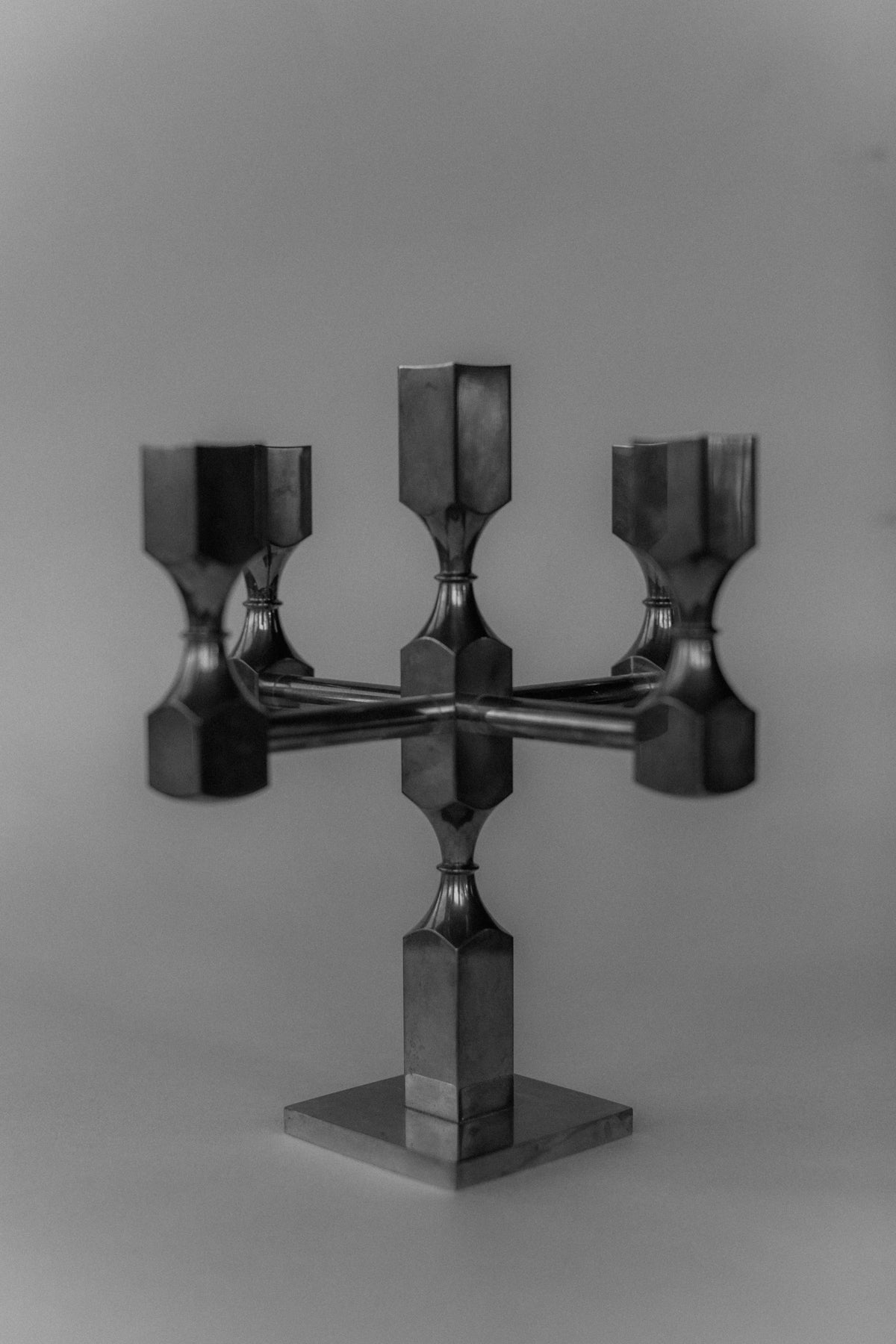 A monochrome image showcases the modern, geometric Brutalist Brass Candleholder by Out For Lunch. This three-arm design, echoing an industrial aesthetic, includes angular shapes and a reflective solid brass surface against a plain background.