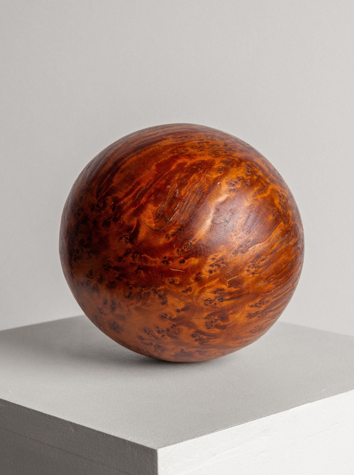 A polished, handcrafted sculpture named the Vintage Burl Wood Spherical Ball Sculpture by Spigel, made of Thuya burl wood with rich reddish-brown hues and intricate grain patterns, rests on a minimalist, light gray pedestal against a plain, soft gray background.
