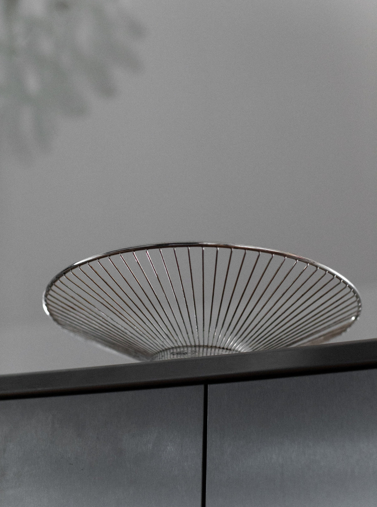 The Out For Lunch String Bowl, a minimalist and modern wire bowl made of metal, sits atop a gray surface with clean lines. Its symmetric design features a series of evenly spaced vertical metal rods radiating outward from a central point at the base, creating an open and airy structure reminiscent of delicate steel strings.