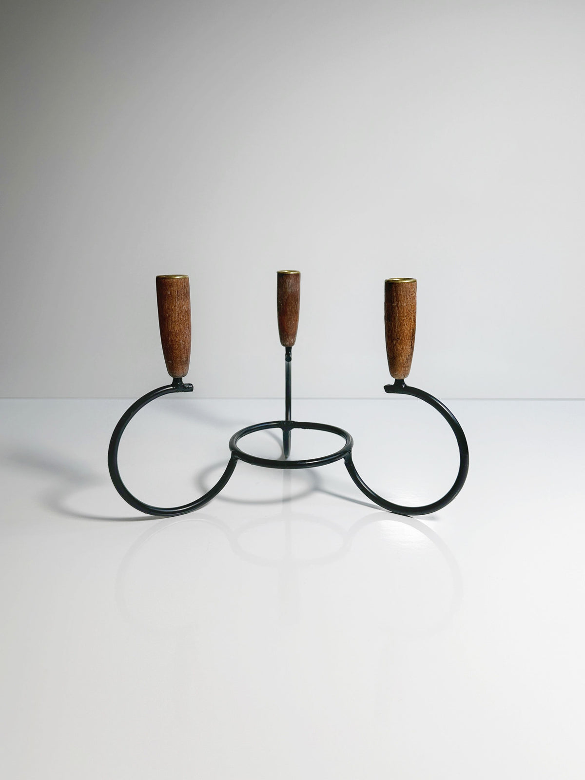 The Danish Candleholder 1960s by Dodo Vintage is a minimalist black metal candle holder that features three circular bases, each topped with teak ends. Designed to hold three candles, it is set against a plain, light background to create a clean and simple aesthetic.