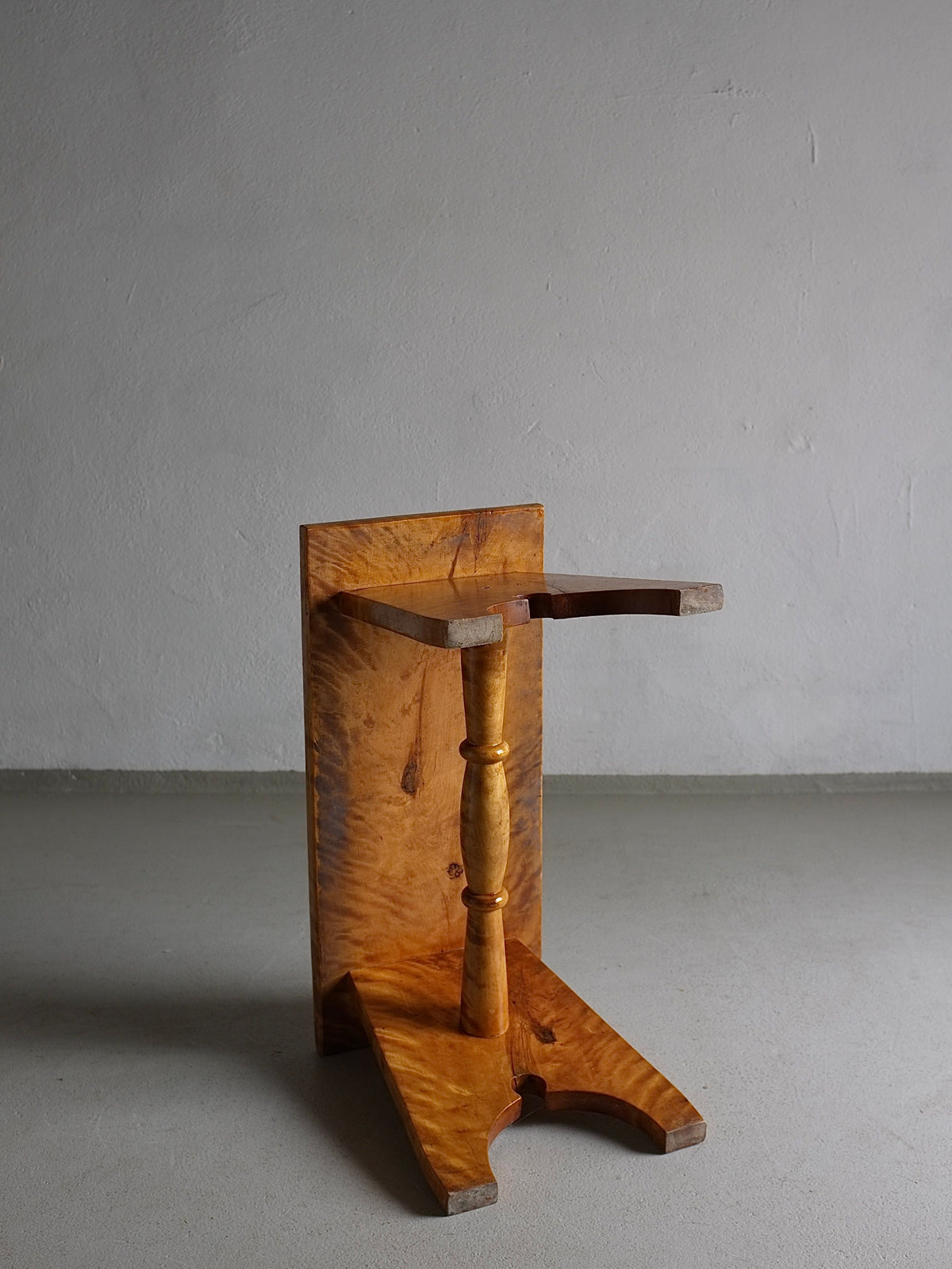 A simple wooden pedestal stand crafted from solid Karelian birch wood with a natural finish. It has a rectangular backboard and a single turned central support connecting a rectangular top and a V-shaped base. The Rustic Solid Karelian Birch Stool | Latvia | 1950s by Veter Vintage is positioned against a plain, gray wall on a concrete floor.
