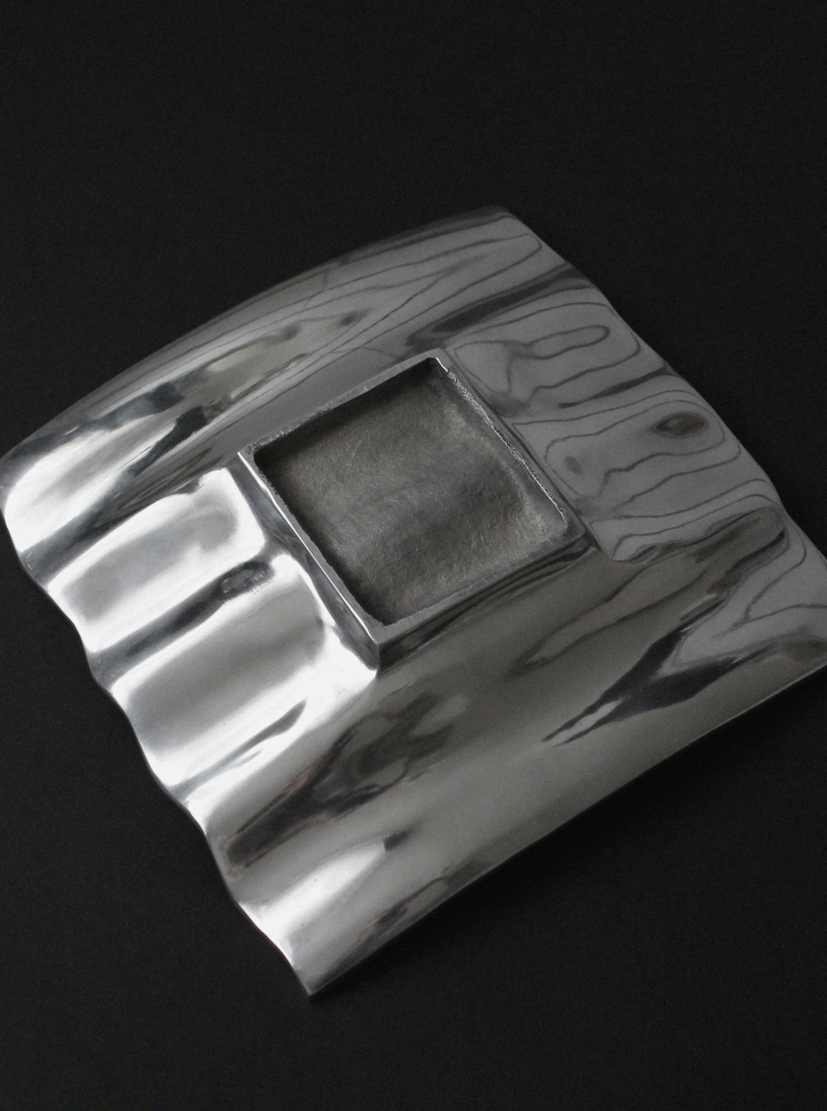 The Polished Aluminum Wave Bowl by Vintage Stories is a square, modern silver brooch featuring a raised rippled surface and recessed textured center, beautifully set against a black background.