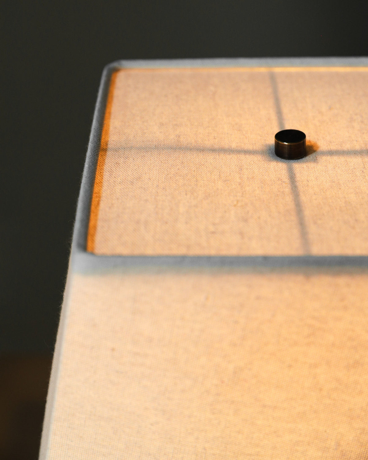 Close-up of the Akune Ceramic Lamp - Number Five by Studio OSKLO, showing its fabric shade and metal finial aglow with warm light, inspired by Japanese artistry, set against a dark, blurred background.