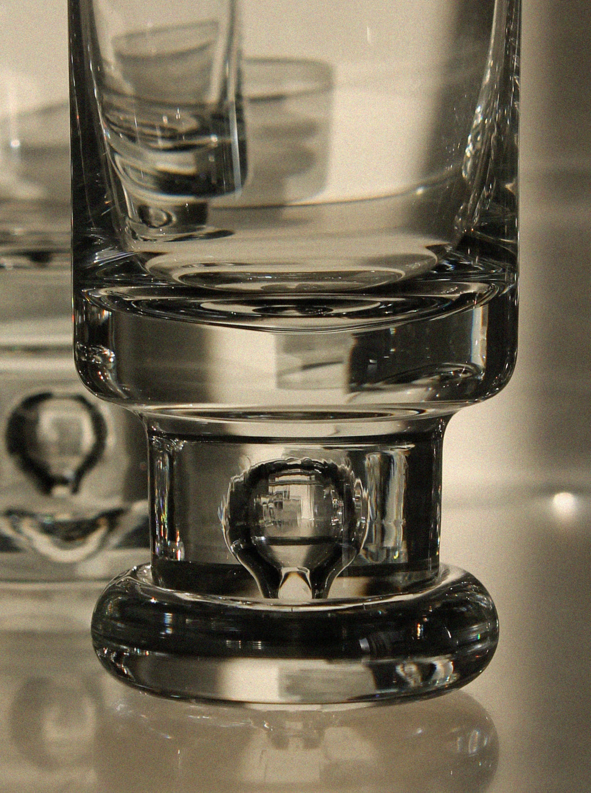 Close-up of the thick, clear glass base from a Vintage Stories Tapio Wirkkala Set of Glasses, showing reflections and distortions in classic mid-century style; other blurred glass objects appear in the background.