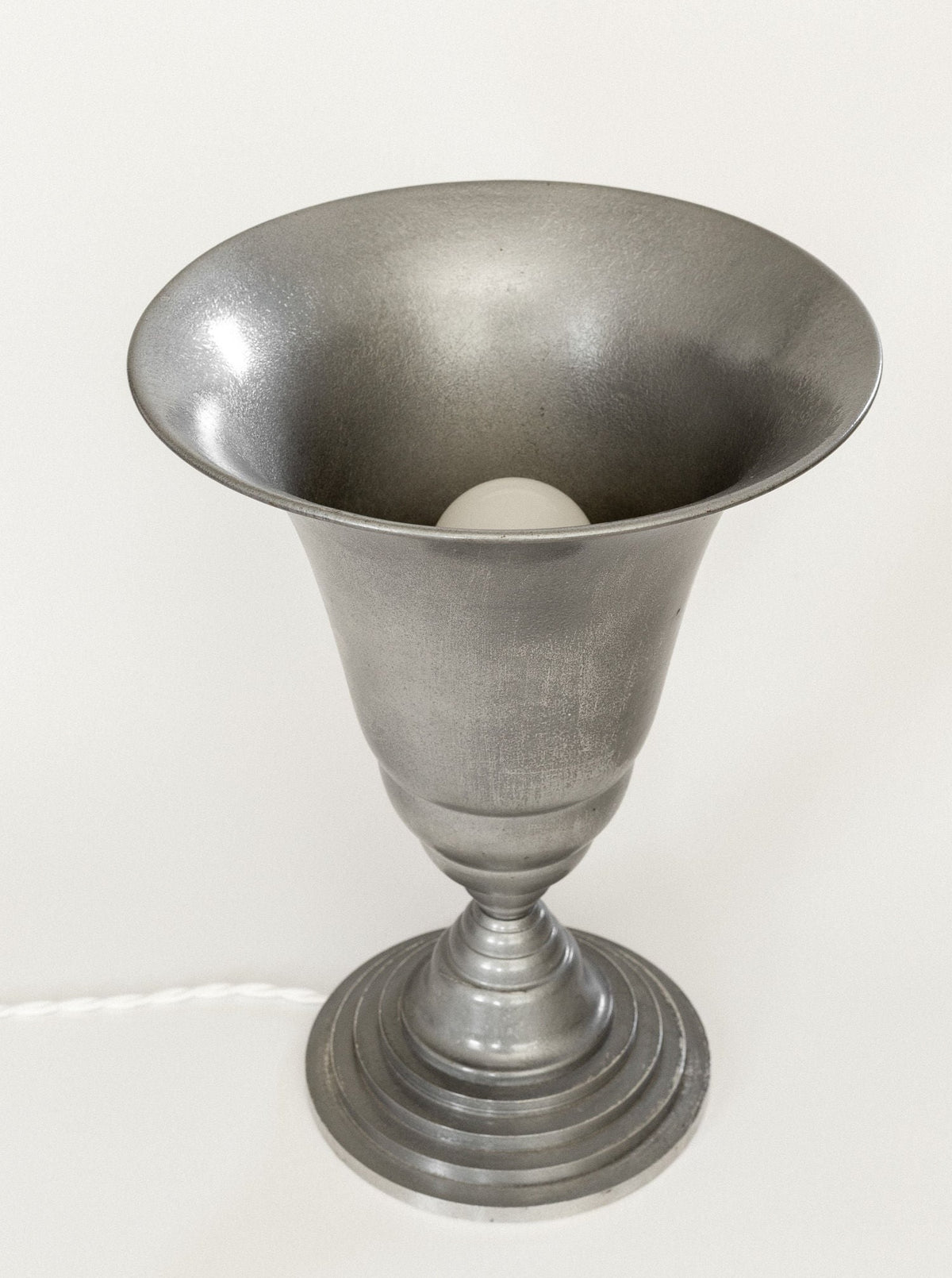 The Art Deco Torchière Table Lamp, France by Spigel is a silver metallic table lamp featuring a conical shape that is wide at the top and narrow at the base, embodying classic French art deco lighting. It has an oxidized aluminum base with a stepped design for a layered appearance, and partially reveals a white light bulb inside. A white power cord extends from its base.