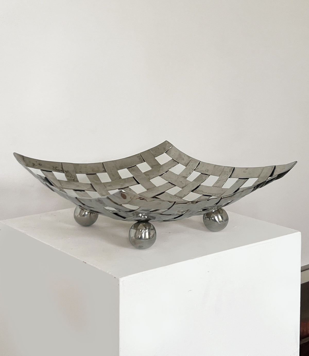 The Porter Studio Aluminum Dish on Feet features a beautiful lattice design and ball feet, elegantly displayed on a white pedestal against a plain wall background.