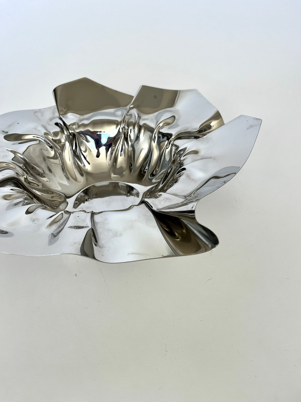 The Folded Chrome Bowl from septembre studios, featuring a shiny, abstract metallic design with wavy, petal-like folds, rests elegantly on a white surface, seamlessly enhancing the interior.