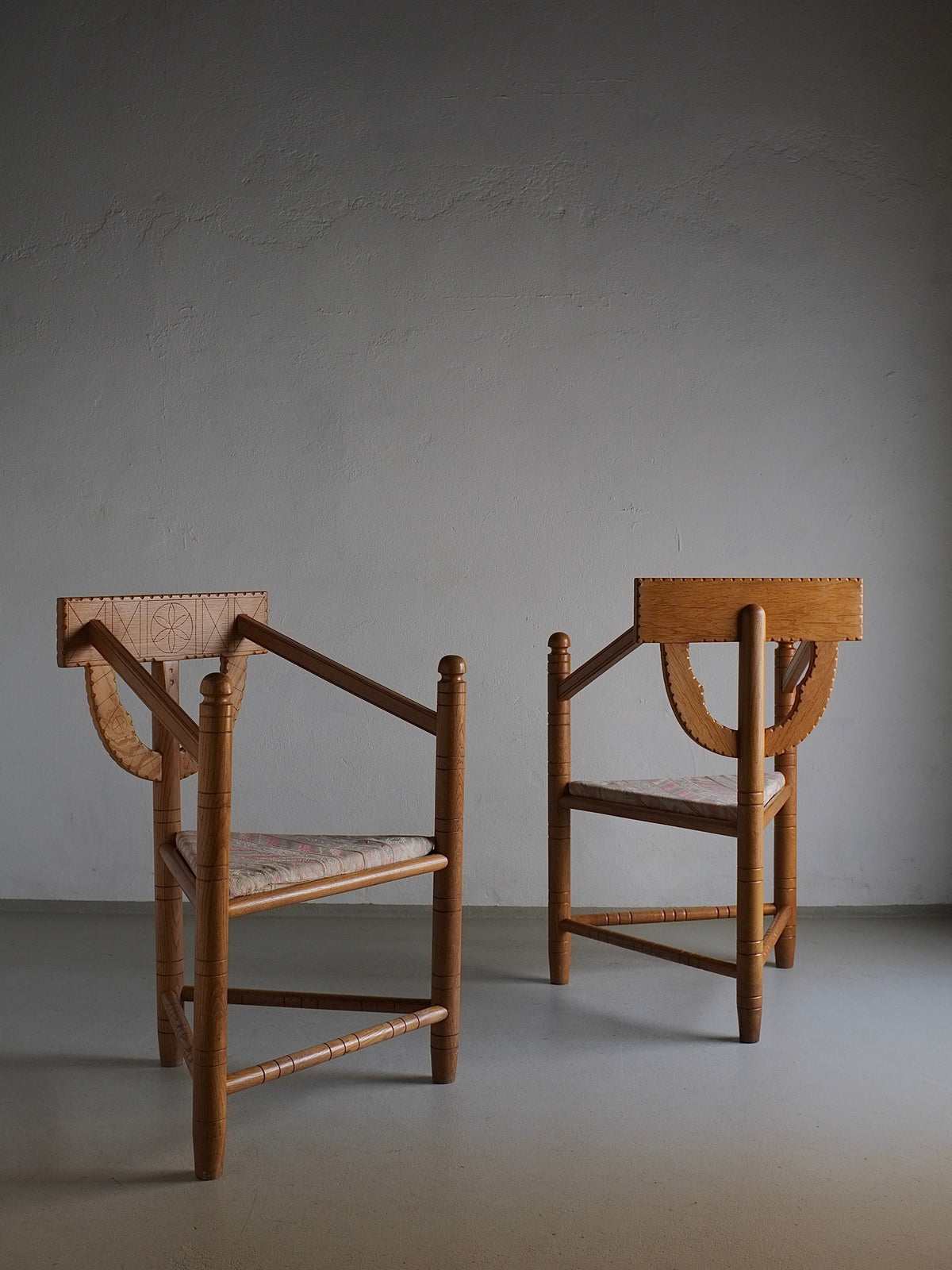 Two intricately designed Rustic Carved Oak Corner Chairs from Veter Vintage, reminiscent of Sweden Design from the 1950s, are positioned facing each other in a minimalist room with a plain, textured gray wall. These chairs feature curved armrests and woven seats, showcasing detailed craftsmanship in vintage condition.