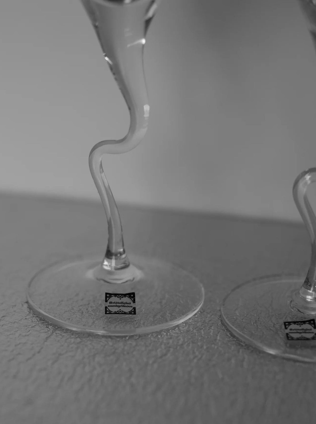 A black and white close-up image of two artisan-made glass stems with curvy designs, resting on a textured surface. Each Set of Vintage Handmade Swirl Champagne Flutes by CU-RA-TED has a logo sticker near the base. The background is blurred, adding to the vintage charm of the scene.