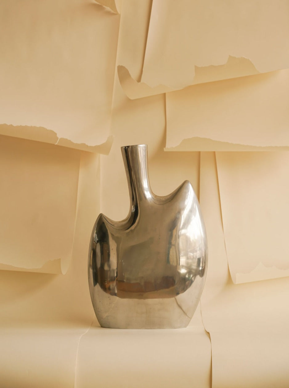 A shiny, polished aluminium Abstract vase XL from Les Objets Amsterdam with a unique broad, asymmetrical shape set against a soft beige background with gently folded paper sheets creating a textured backdrop.