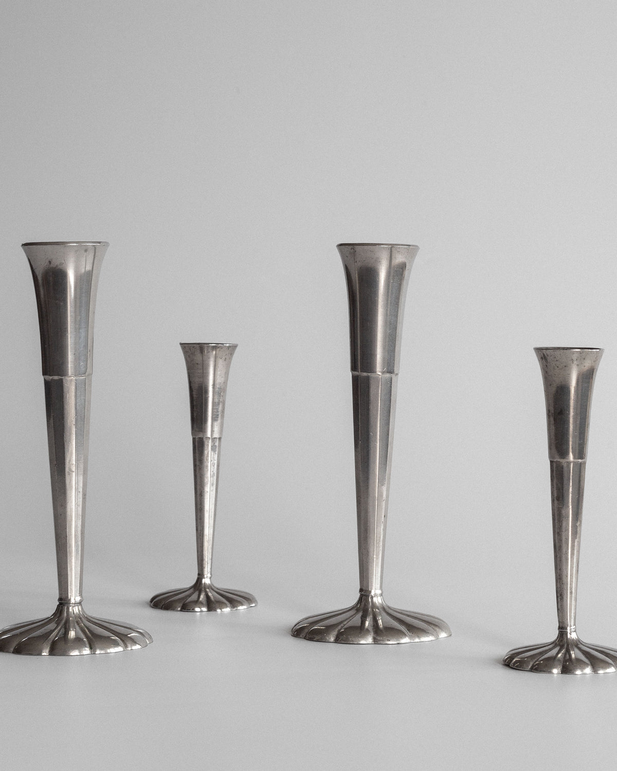 A set of four sleek, metallic Spigel vases, varying in height and featuring elongated designs, stand on a plain surface with a neutral background. Reminiscent of Scandinavian style and echoing Just Andersen's Art Deco influence, they boast modern minimalism with reflective surfaces.