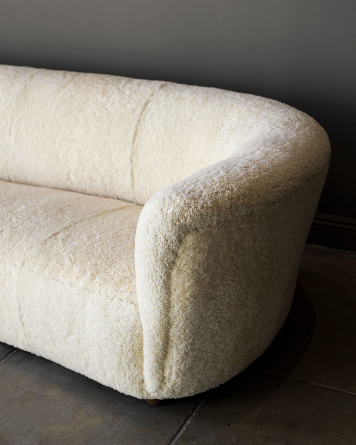 The Fritz Hansen Sofa in Shearling by Studio OSKLO features soft, cream-colored upholstery with a shearling texture, rounded edges, and sits on a tiled floor against a gray wall.