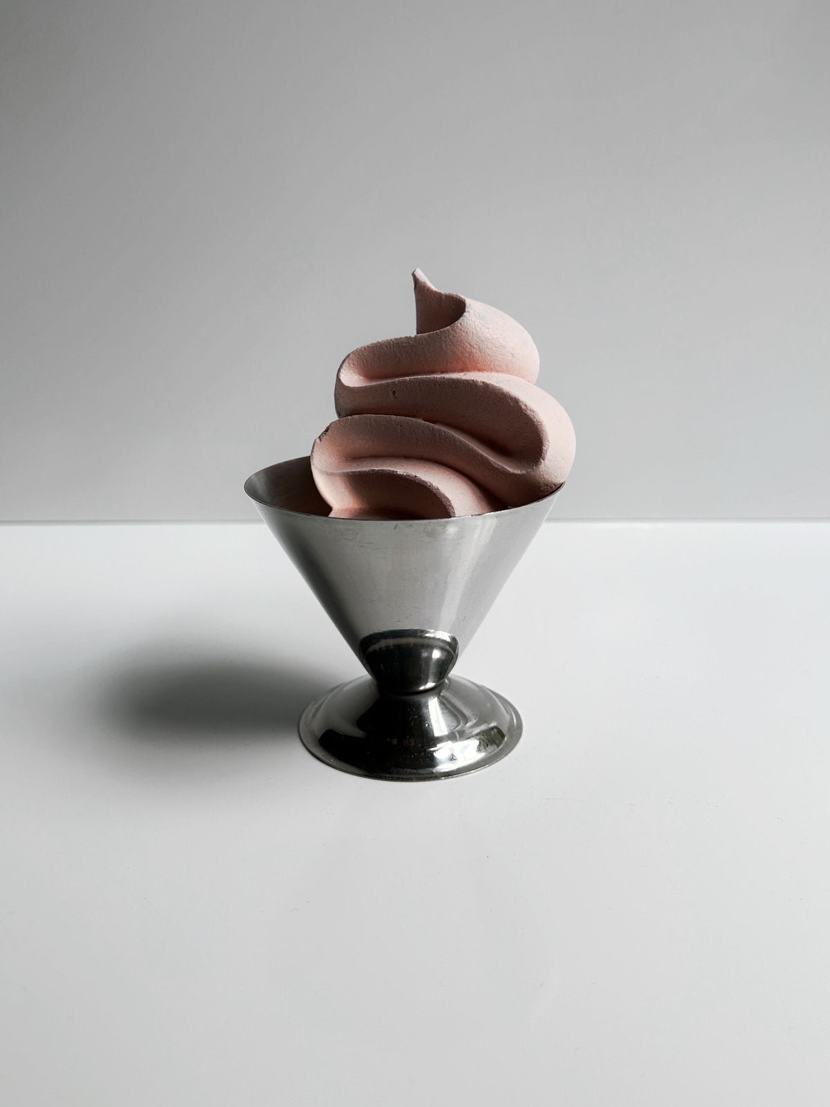 A soft serve ice cream with a light pink hue, evoking the vintage charm of the 1980s, nestled in a shiny Italian Stainless Steel Dessert Cup from Dodo Vintage against a plain, light gray background.