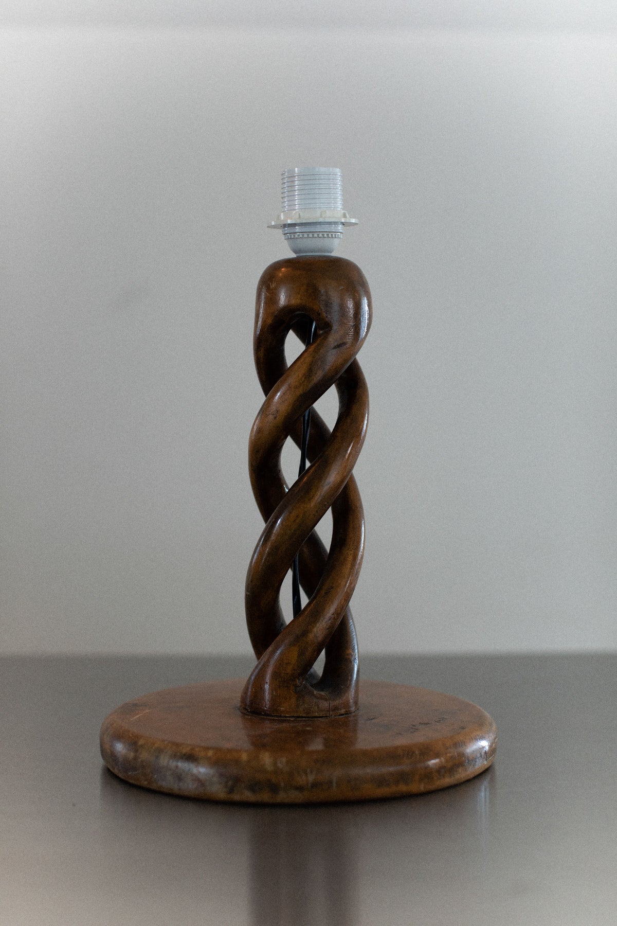 The Early-Century Lamp by Out For Lunch showcases a twisted wooden base with an intricate intertwining spiral design, elegantly positioned on a flat surface against a neutral background. The double helix shape leads to a top socket, ready for a light bulb.