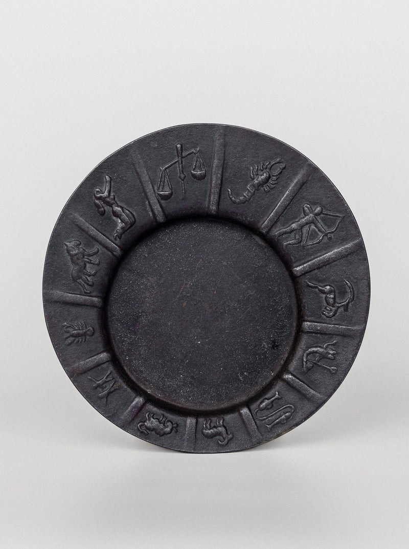 The "Art Deco Zodiac Signs Decorative Dish" by Spigel is a round, dark-colored plate featuring the 12 astrological zodiac symbols embossed along the rim. This Scandinavian vintage design showcases equally spaced symbols starting from Aries and ending with Pisces, arranged clockwise around the center of the dish.