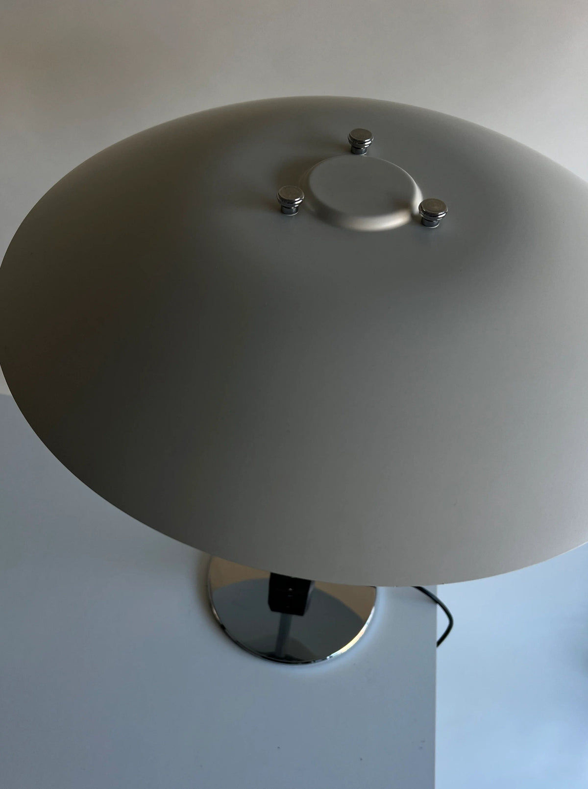 A close-up shot of a modern table lamp with a sleek, gray metal shade and a chrome base. The CU-RA-TED Poulsen Table Lamp PH 4/3 1983, reminiscent of Poul Henningsen's 1983 designs, features three small screws on the top of the shade. The background and surface are both gray, creating a minimalist aesthetic.