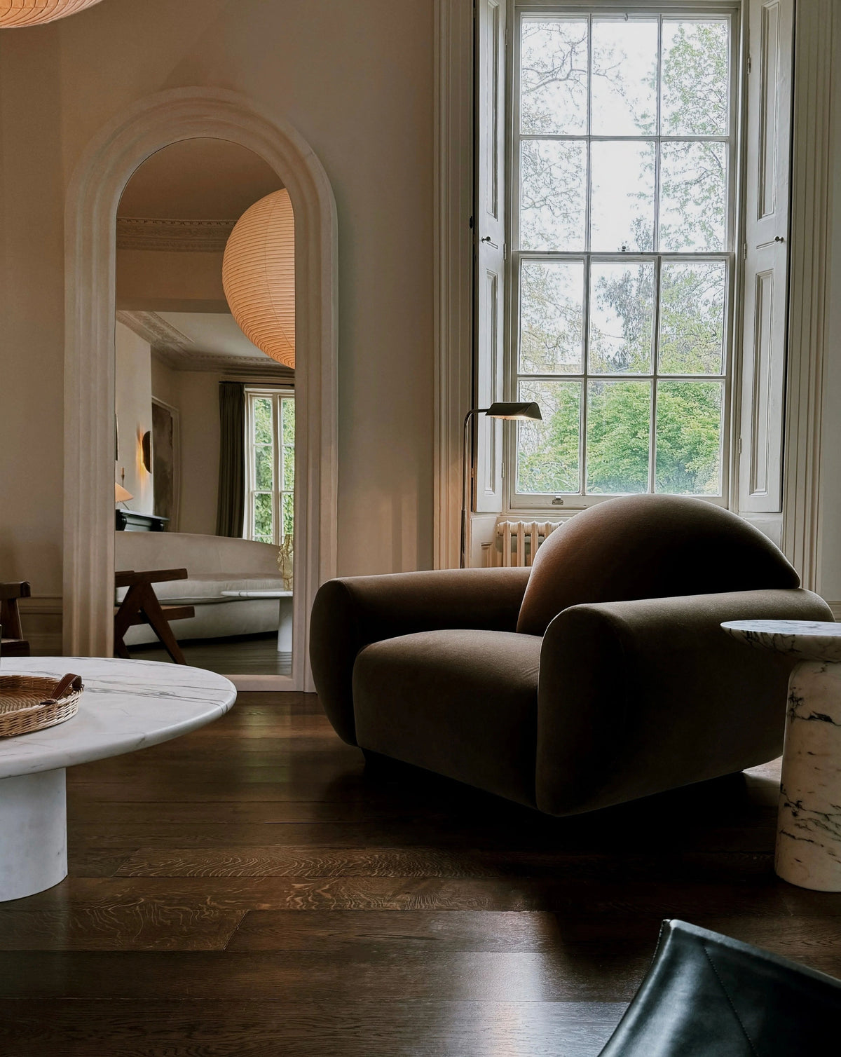 In a cozy living room, you’ll find the "Sparta" armchair by Ransom & Dunn, elegantly draped in alpaca wool. A rounded marble coffee table sits nearby alongside a tall lamp beside a large window showcasing lush greenery. A mirror enhances the room's depth and warmth, completing the elegant atmosphere.