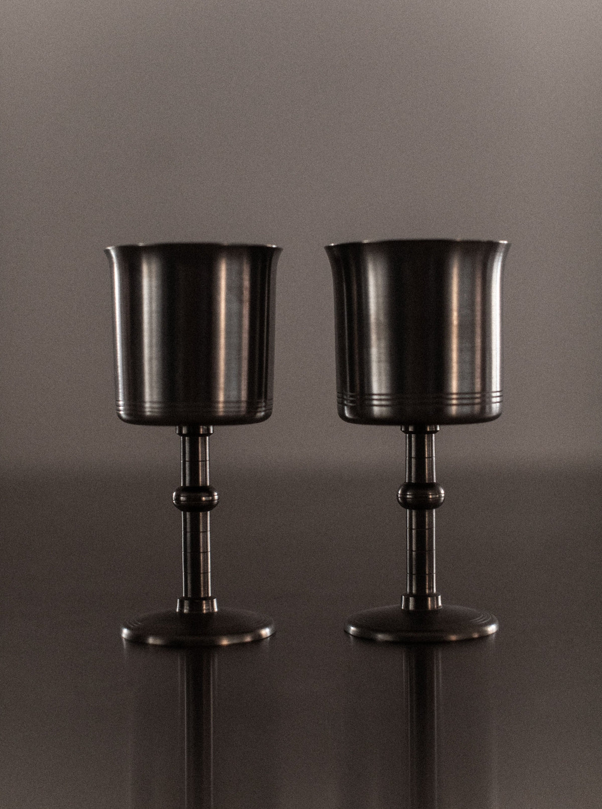 Two Art Deco Cups from Out For Lunch, with smooth and reflective surfaces, stand side by side on a glossy surface, creating a symmetrical arrangement. The background is neutral and softly lit, adding to the timeless elegance and classic appearance of the scene.
