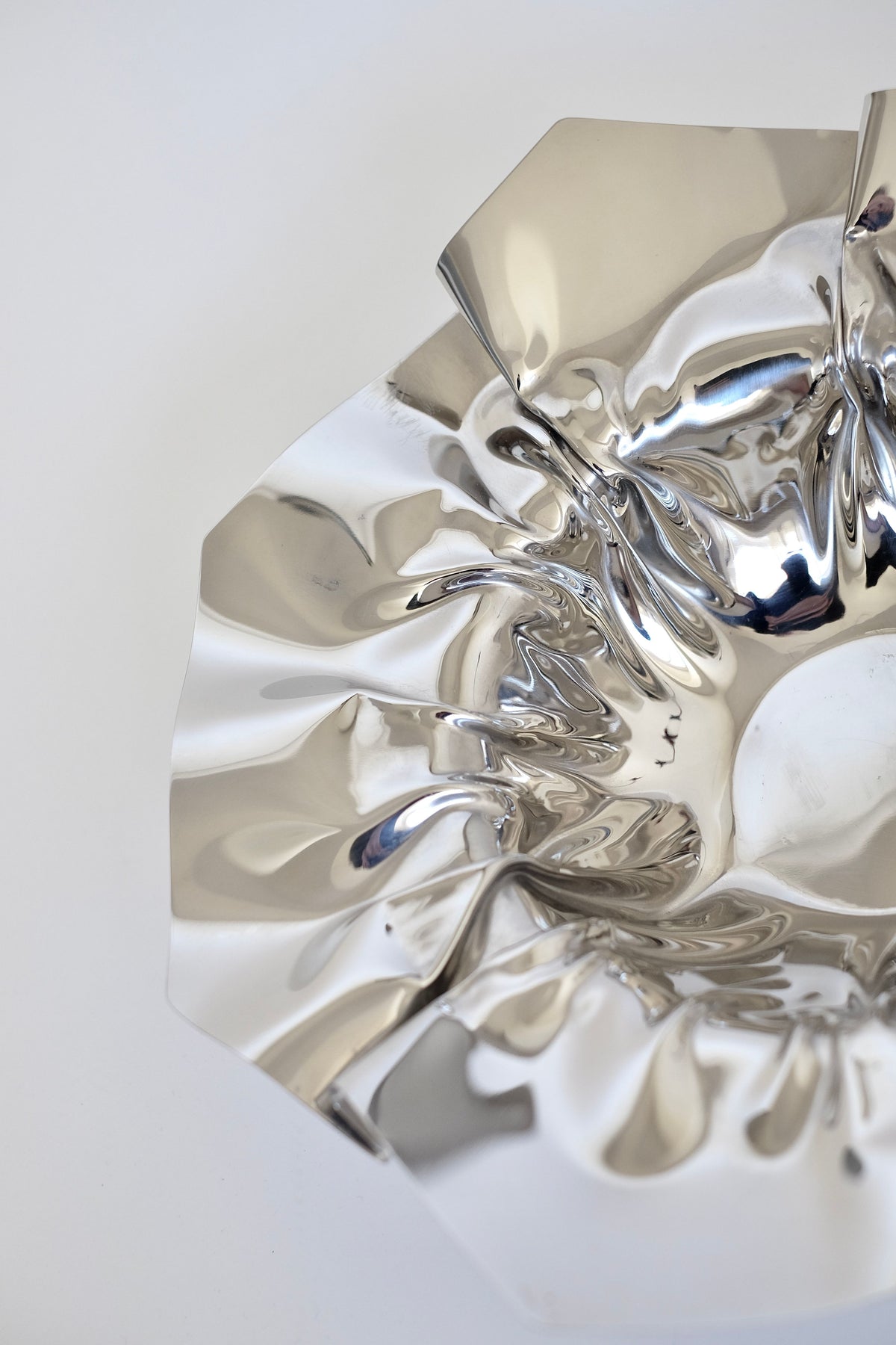 A close-up of the Folded Chrome Bowl by septembre studios showcases its shiny, metallic surface with a wavy, reflective design and an intriguing folded shape. The smooth interior captures light beautifully, creating intricate patterns and highlights against a plain, light-colored background.