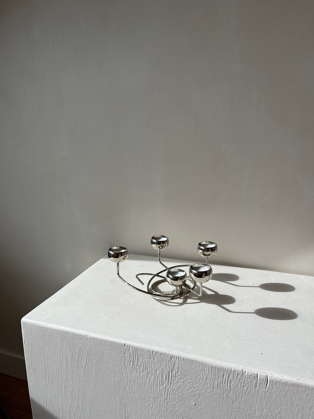 A contemporary "Chrome Candleholder" from septembre studios, featuring five circular holders, is elegantly displayed on a minimalist white pedestal against a plain wall. Its vintage-inspired design highlights distinctive angles as soft natural light creates refined shadows across the surface.