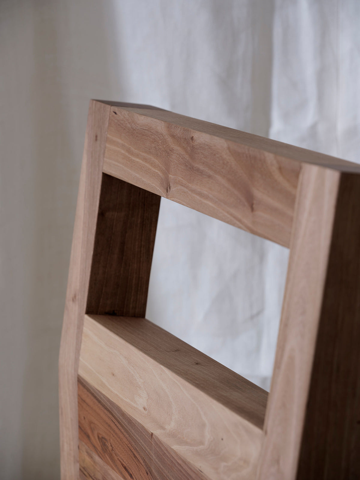Close-up of the "Forma" Armchair backrest from FEBRERO SHOP, crafted in solid light-brown wood with a central rectangular cutout. The rich grain texture beautifully contrasts with softly blurred linen fabric in the background.