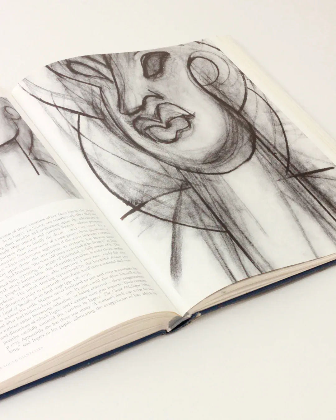 An Henri Matisse Vintage Book from Boga Avante Shop lies open on a white surface, revealing a charcoal sketch of an abstract face with exaggerated features, reminiscent of modern art. Text adorns the lower part of the left page, partially visible but not entirely readable.