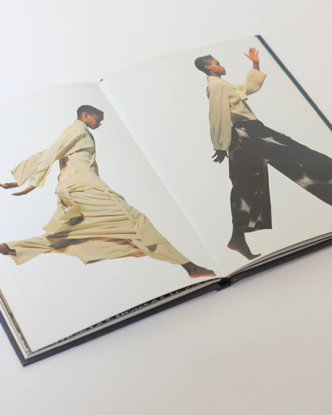 The Issey Miyake Book from Boga Avante Shop presents an open book view featuring two images of a person in dynamic poses. On the left page, it highlights Issey Miyake's iconic pleating techniques with a full-length flowing outfit captured mid-leap. Meanwhile, the right page depicts them confidently stepping forward in contemporary fashion, wearing wide-leg pants and a loose top.