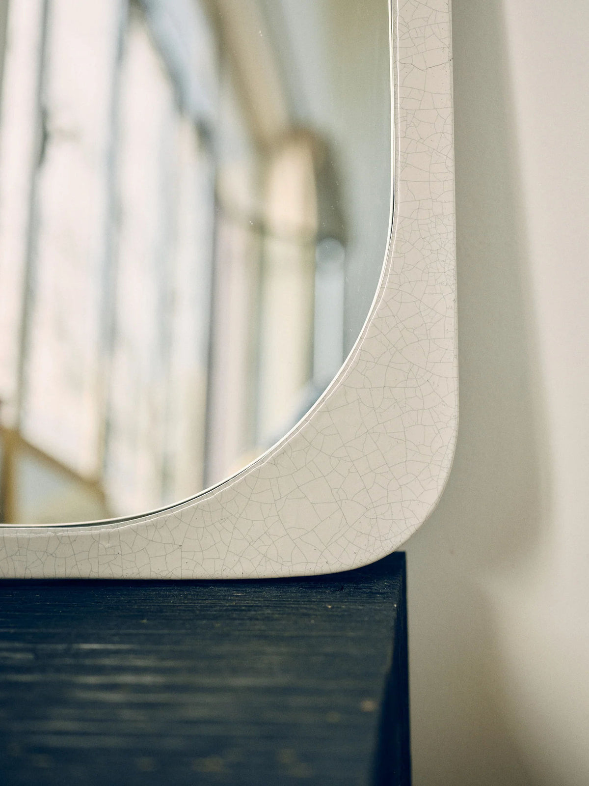 A close-up of a cracked white ceramic surface with a crackled enamel finish reveals fine lines and a corner with curved edges. The Mirror Bien Fait in Lava Stone by Bien Paris, made in Italy, rests on a dark wooden shelf, with blurry reflections of a window and light in the background.
