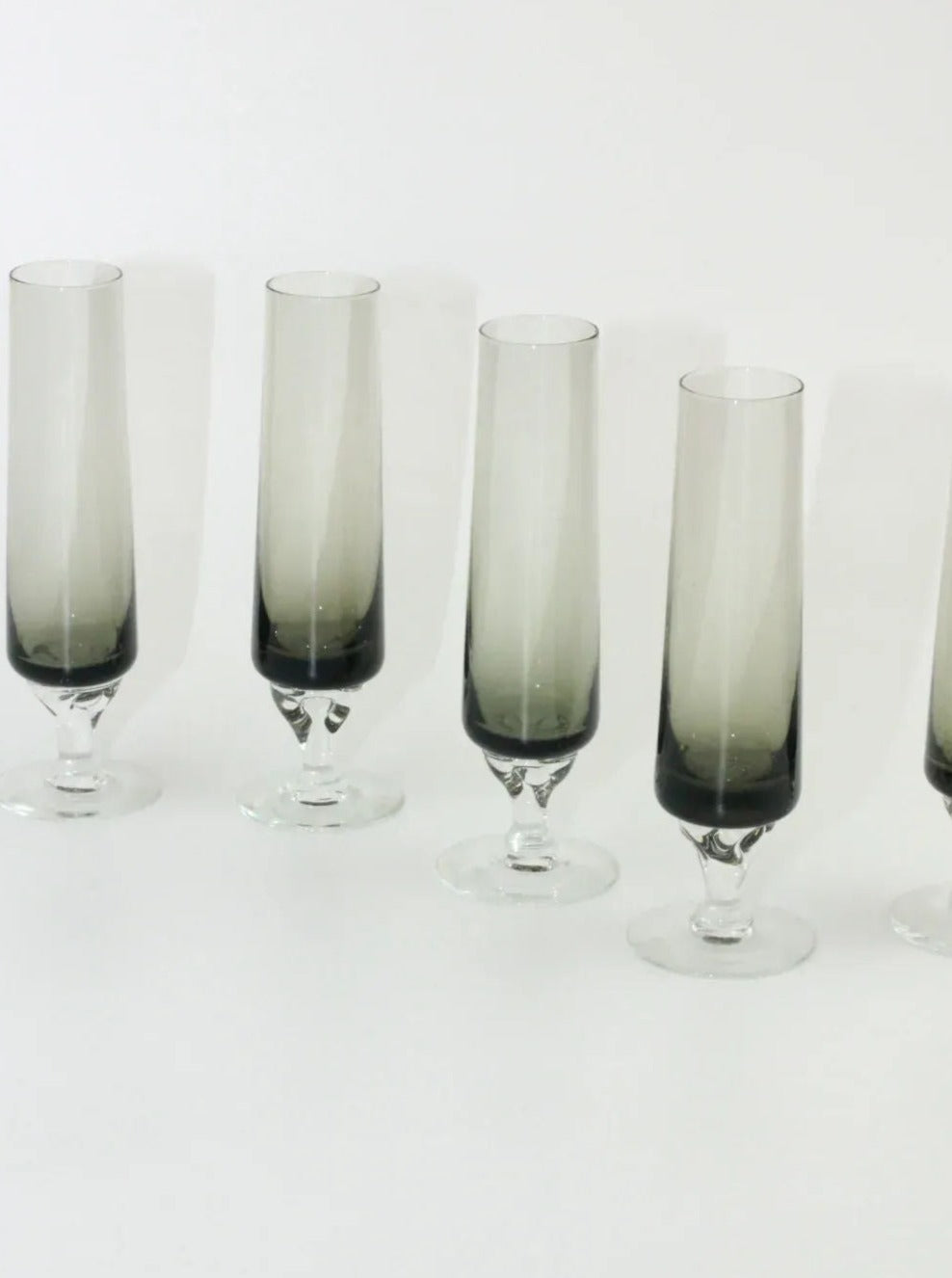 Five slender, gradient-shaded champagne glasses with smoky gray stems and clear tops stand in a row on a white surface. The Boga Avante Shop Set of Five Vintage Flute glasses boast a modern and minimalist design, emphasizing the smooth transition between the dark and clear glass sections.