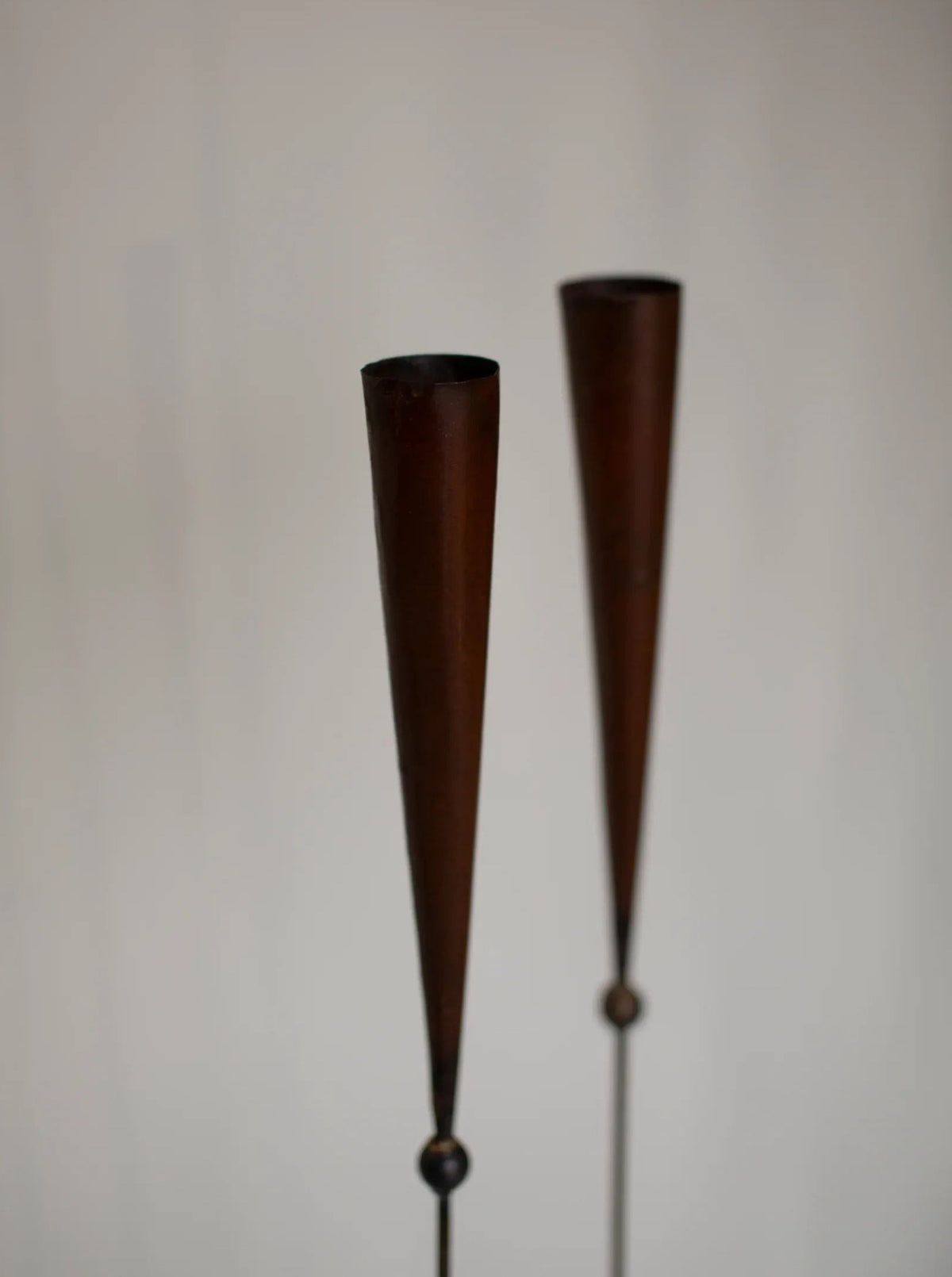 A close-up of two vertical, tapered wooden objects with a conical shape, resembling handmade candle holders or decorative spindles, set against a plain, light background. These 80's signed pieces by Pieter Spruyt have a dark brown finish and are minimalist in design from the CU-RA-TED brand appear.