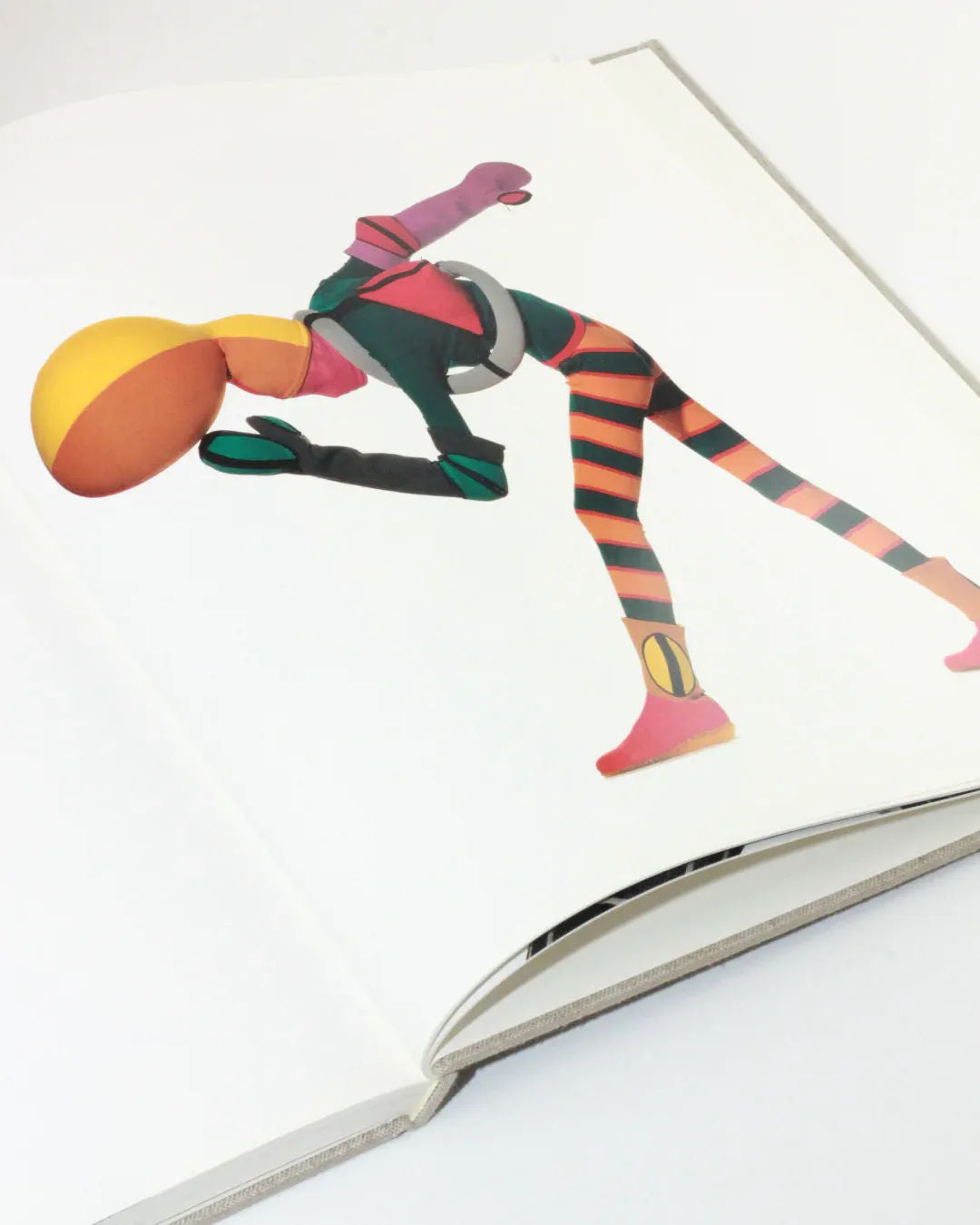 An open book, "Irving Penn regards the work of Issey Miyake" from Boga Avante Shop, showcases an abstract, colorful figure in a dynamic pose that evokes the innovative fashion designs of Issey Miyake. The figure features geometric shapes for its head and body, adorned in vibrant yellow, orange, and green with striped leggings against a plain white background.
