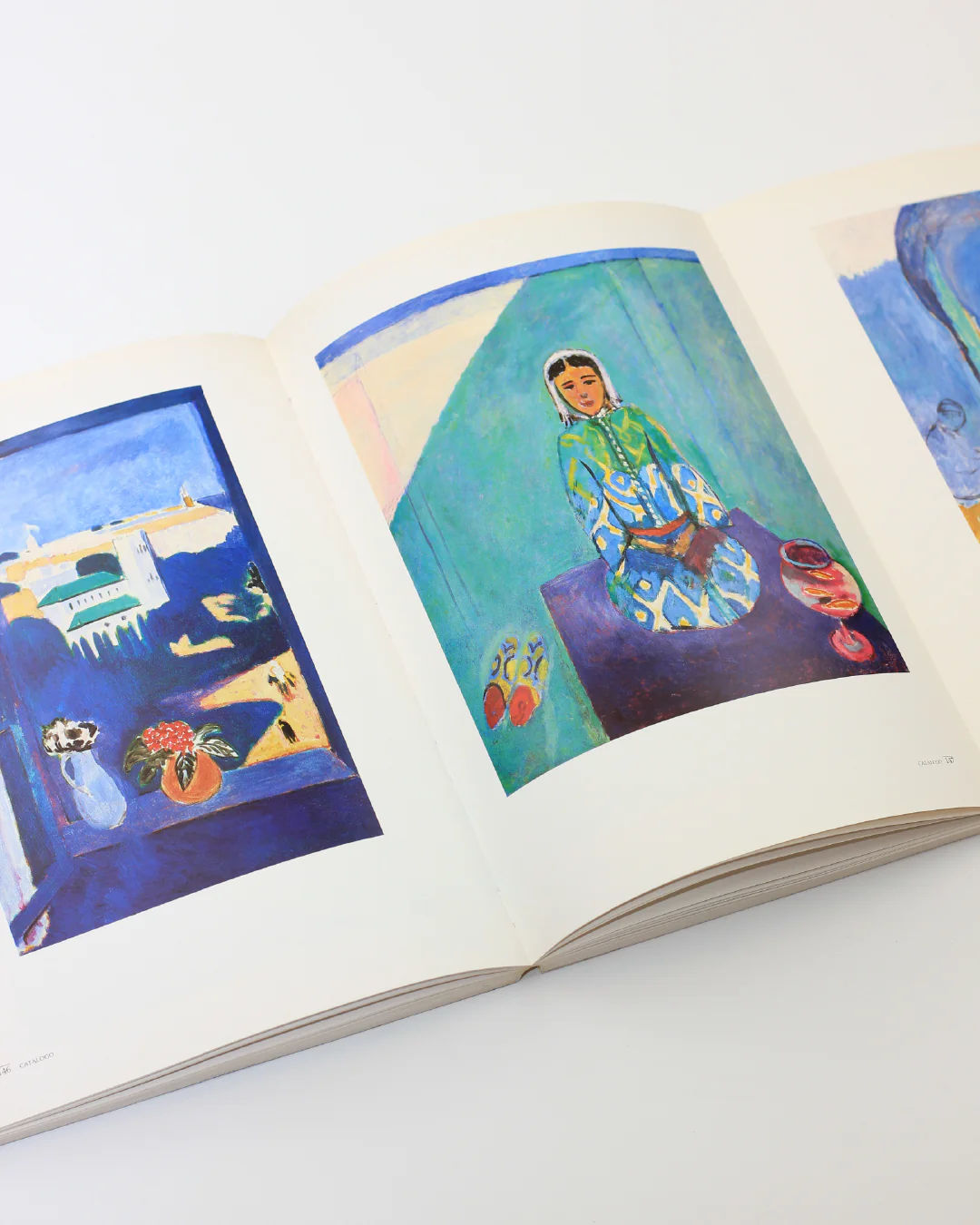 An open Henri Matisse Book from Boga Avante Shop reveals colorful pages. On the left, there's a painting with blue tones and flowers in a vase, reminiscent of Fauvism. The right page showcases an individual in patterned clothing set against a vibrant blue and green background, reflecting the iconic modern art style of Henri Matisse.