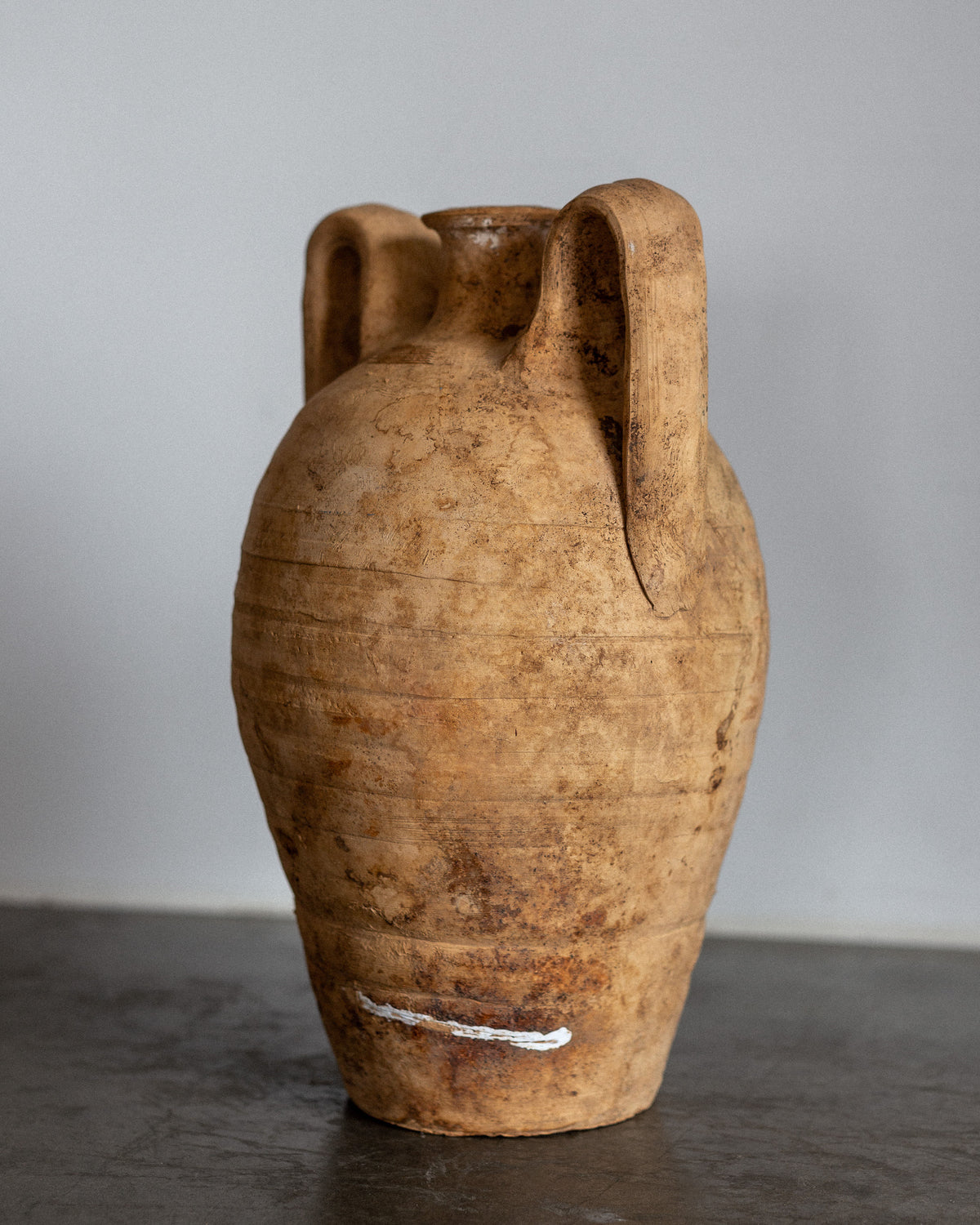 The Early 20th Century European Ceramic Amphora by Spigel is a rustic, tall earthenware piece with two looped handles. It rests on a gray surface and features a weathered, earthy texture in brownish-tan with visible horizontal grooves and a small white mark near the base.