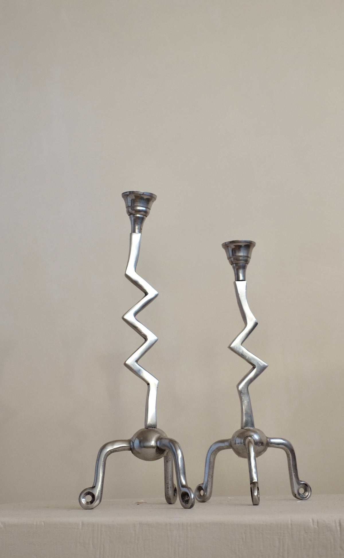 A pair of Zigzag Candleholders from septembre studios, featuring zigzag stems and tripod bases, stand side by side against a plain background. Their distinctive design brings an artistic flair to the setting, making them a genuine zilver treasure.
