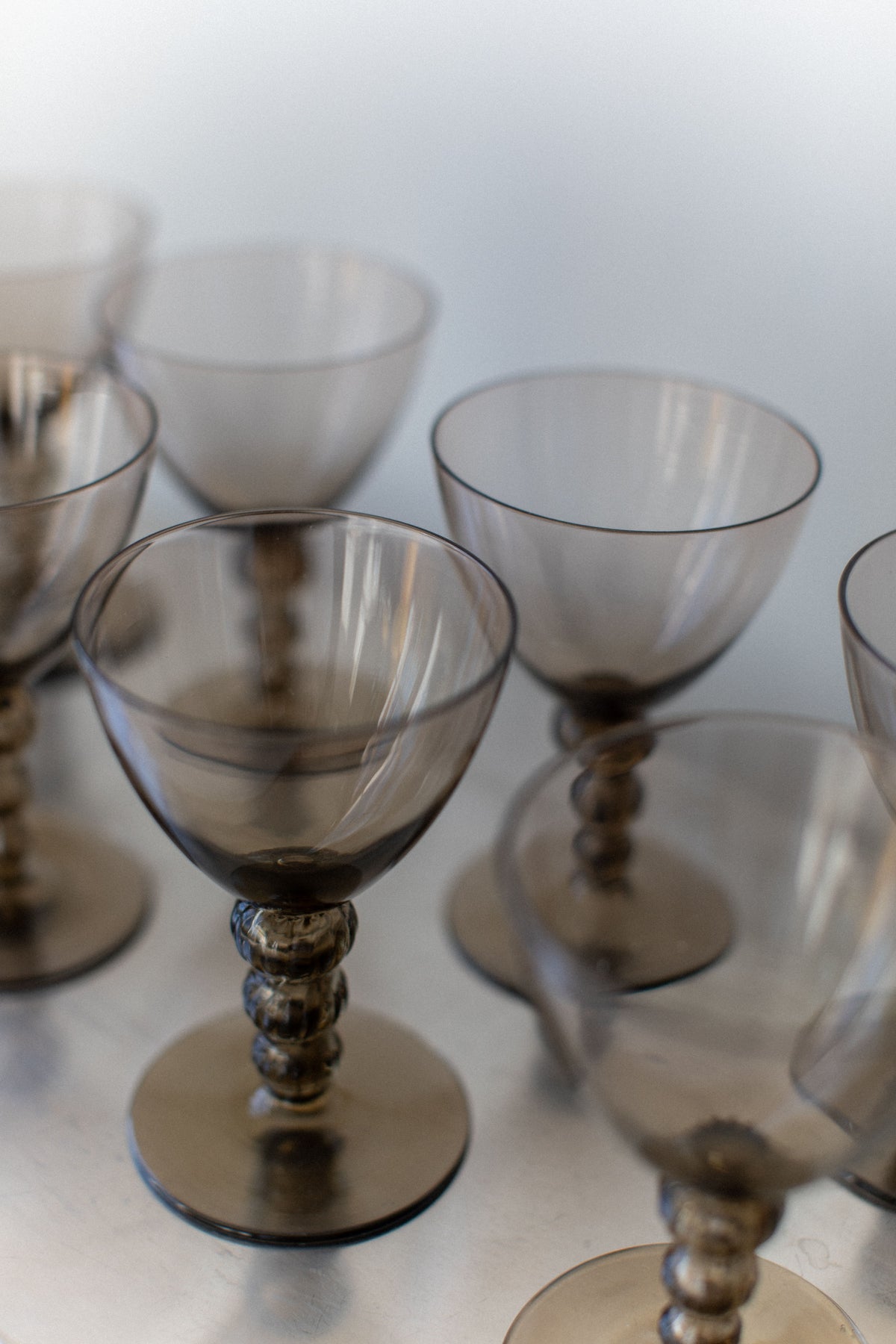 The Art Deco Glass collection by Out For Lunch, featuring translucent gray goblets with ornate, twisted stems, elevates the sophistication of any drinking experience. Arranged elegantly on a reflective surface, these glasses cast soft reflections and shadows, narrating a unique natural story.