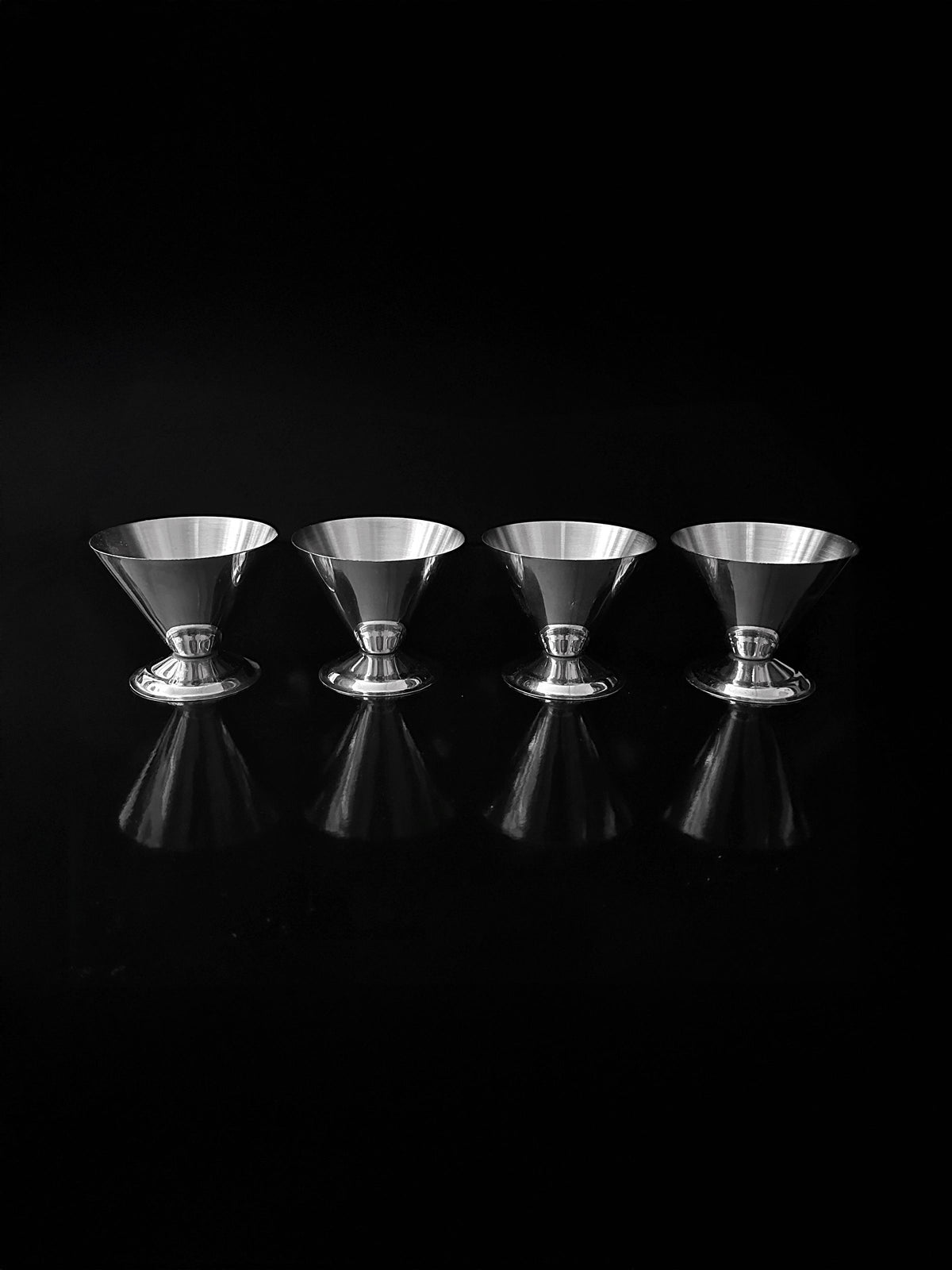 A set of four Italian stainless steel dessert cups by Dodo Vintage is arranged in a row against a black background. Their reflective surface creates symmetrical reflections beneath each cup, accentuating their sleek design reminiscent of elegant 1980s Italian martini glasses.