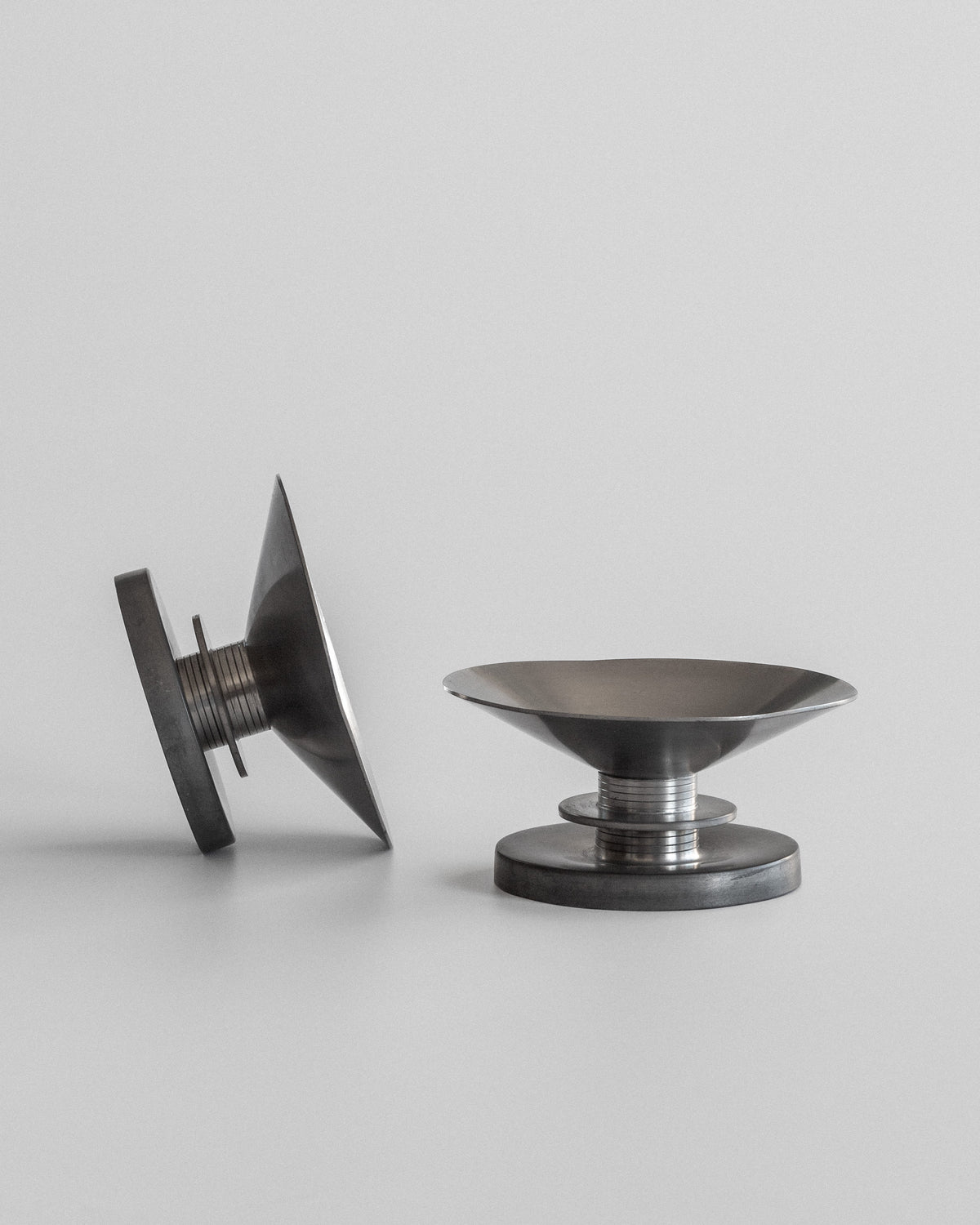 The Spigel Modernist Candleholders by Sylvia Stave from the 1930s are metallic objects displayed against a plain background; one stands upright like a wide-based shallow bowl, while the other lies showcasing a threaded section, exemplifying sleek industrial design and 20th-century decorative arts.