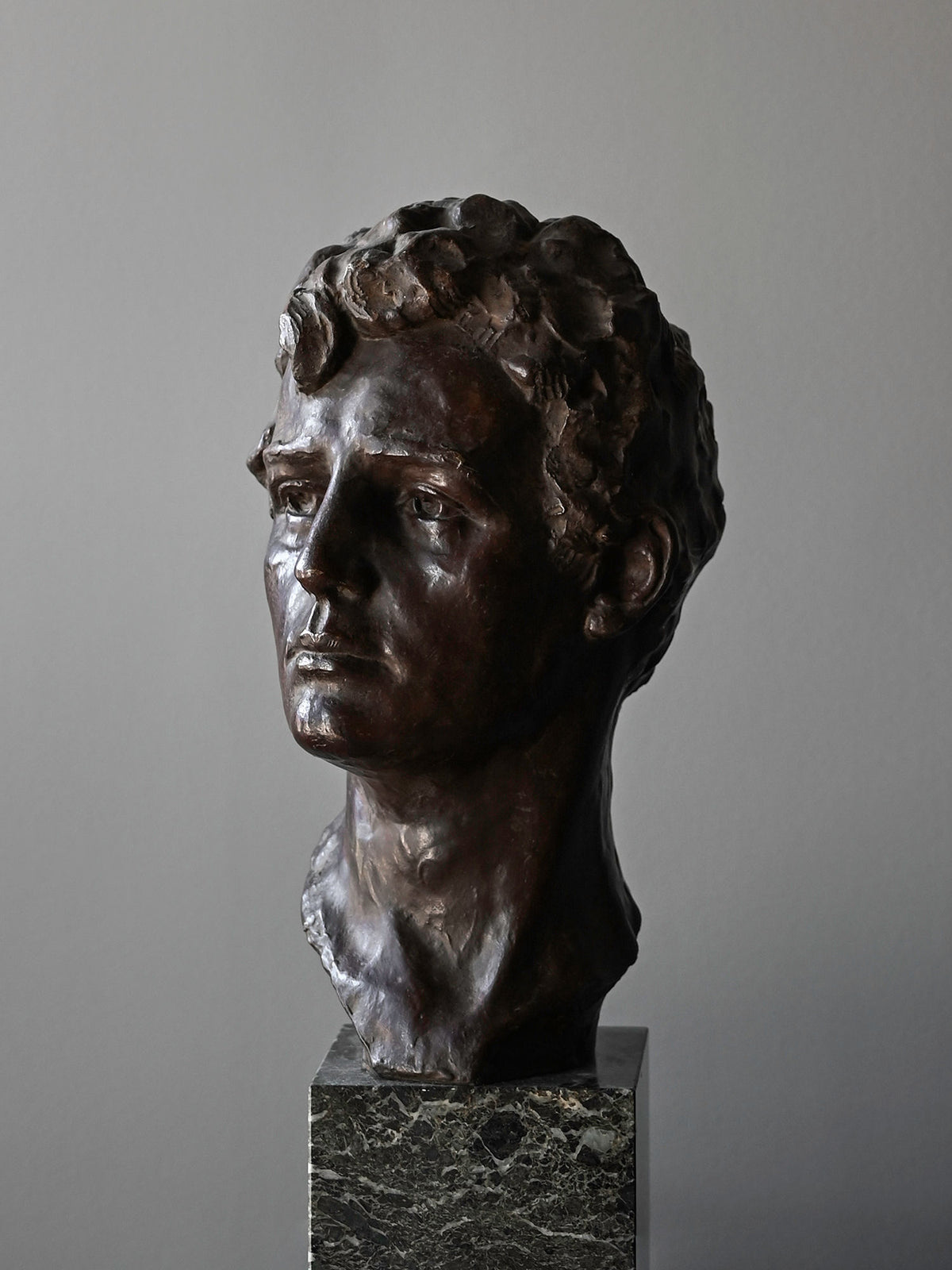 The Bronze Bust of Male by Victor Demanet from Studio OSKLO features wavy hair and lifelike detail, set on a dark marble pedestal and shown against a plain gray background, highlighting Belgian bronze artistry.