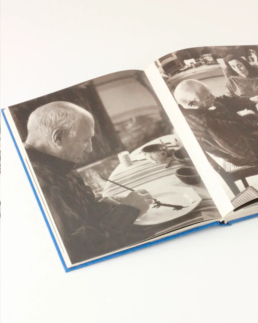 An open book displays black-and-white photos of an elderly man painting with a brush on a circular canvas. An intimate portrait by Boga Avante Shop, the images of Picasso and Jacqueline capture the artist's deep focus, while a young woman named Jacqueline is visible in the background on the right page.
