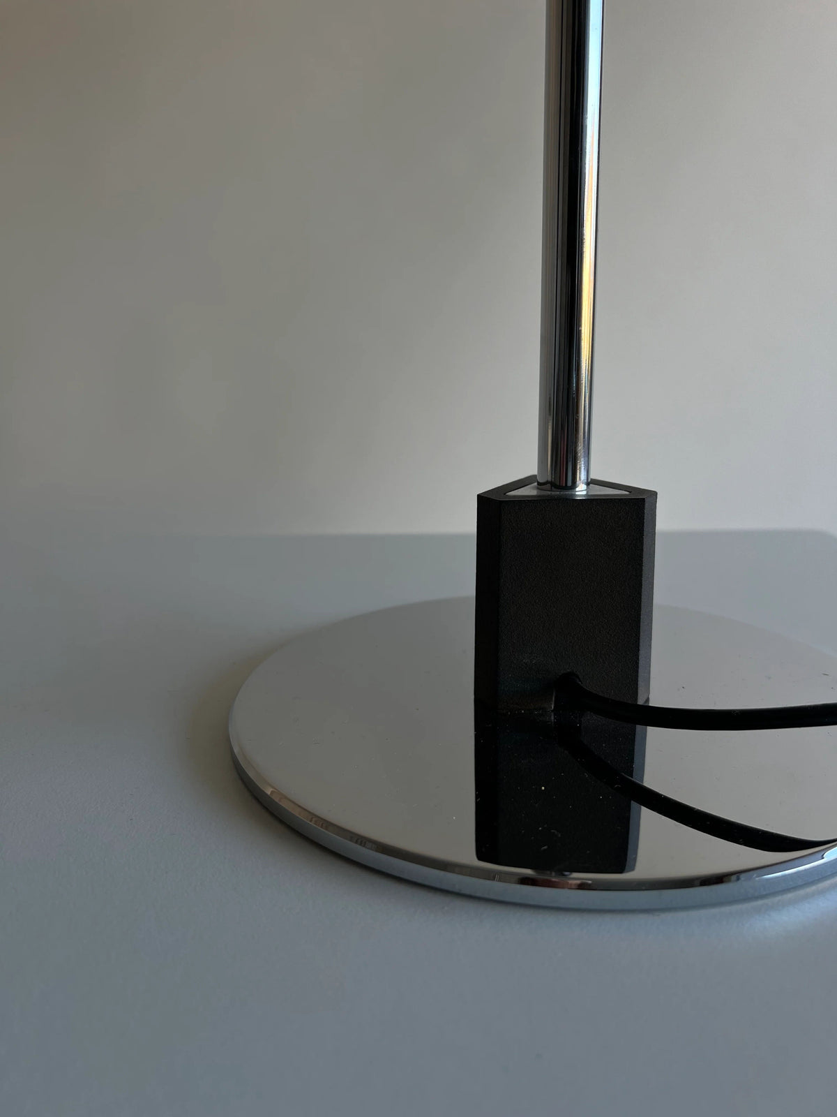 Close-up of a modern CU-RA-TED Poulsen Table Lamp PH 4/3 1983 base with a sleek, reflective chrome finish. The black cord is plugged into a rectangular block at the base, and the lamp stem extends vertically from the center of the circular base, reminiscent of Poul Henningsen’s vintage table lamp designs. The background is plain and out of focus.