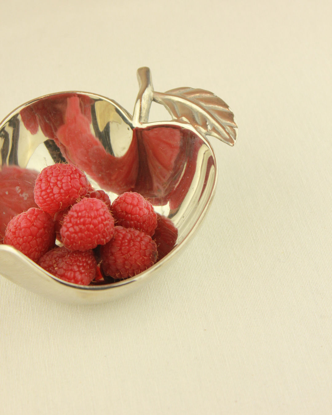 The Decorative Apple Shaped Dish by Boga Avante Shop, crafted in silver with a leaf detail, displays fresh raspberries on a plain, light beige background.