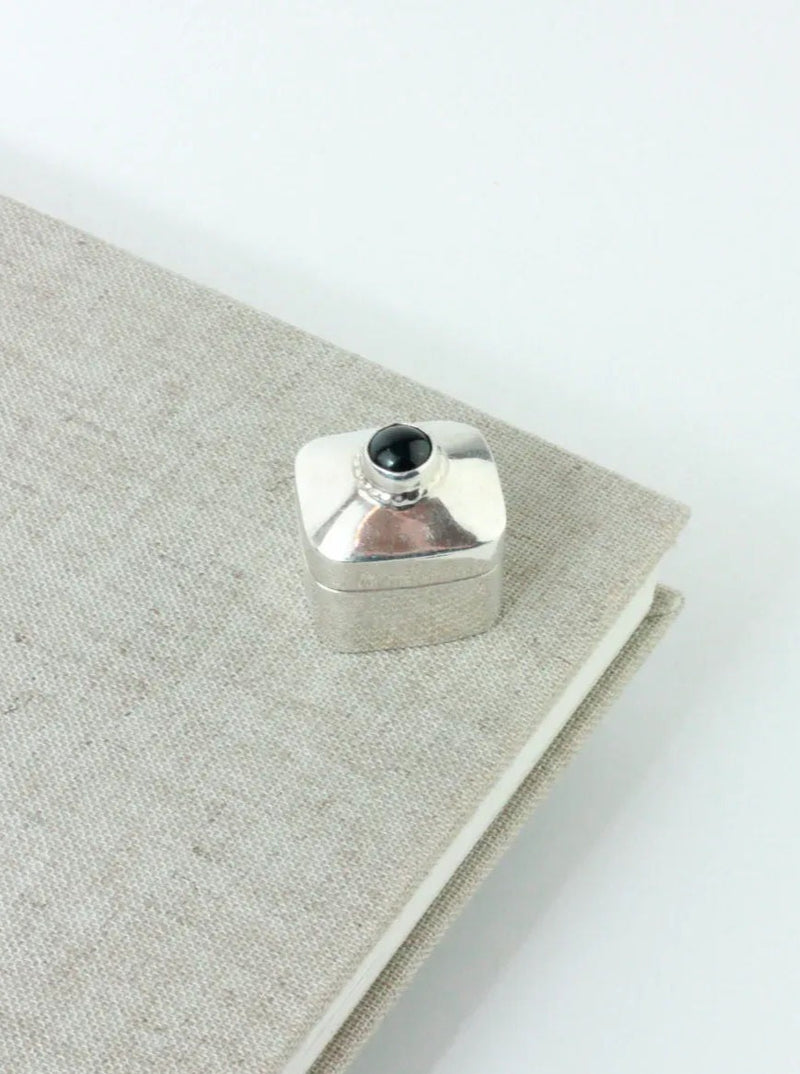 A small, square, silver-colored Silver Pillbox With Onyx Stone by Boga Avante Shop with a rounded top sits on the corner of a closed, textured, beige notebook. This handmade Taxco Mexico piece features a black circular onyx stone on top. The background is plain and white.